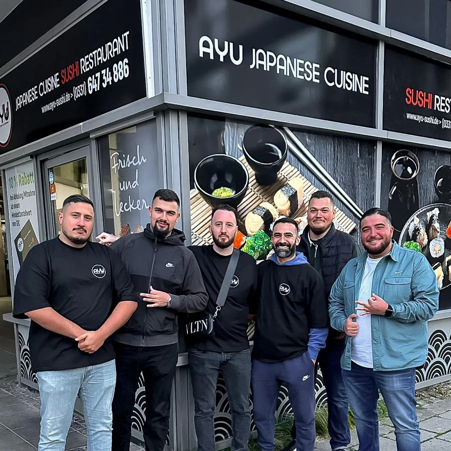 Sechs Männer vor dem Restaurant Ayu Japanese Cuisine, über ihnen der Schriftzug, hinter ihnen Schaufenster mit Sushi-Motiven. Dritter von links: Inhaber Oguz Han Celik im schwarzen Ayu-Shirt mit Umhängetasche.